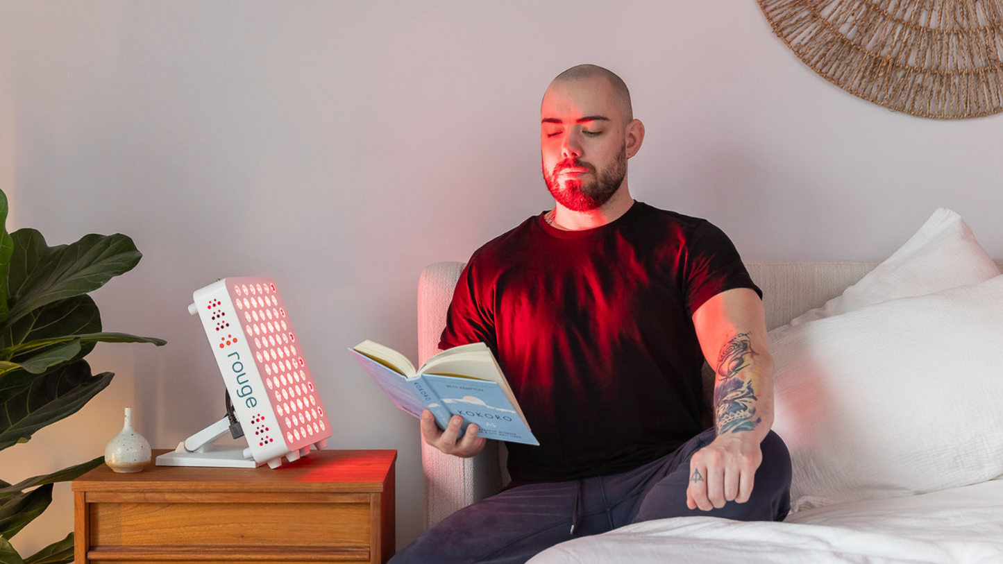 A men sitting on the bed and using red light therapy, while reading a book.